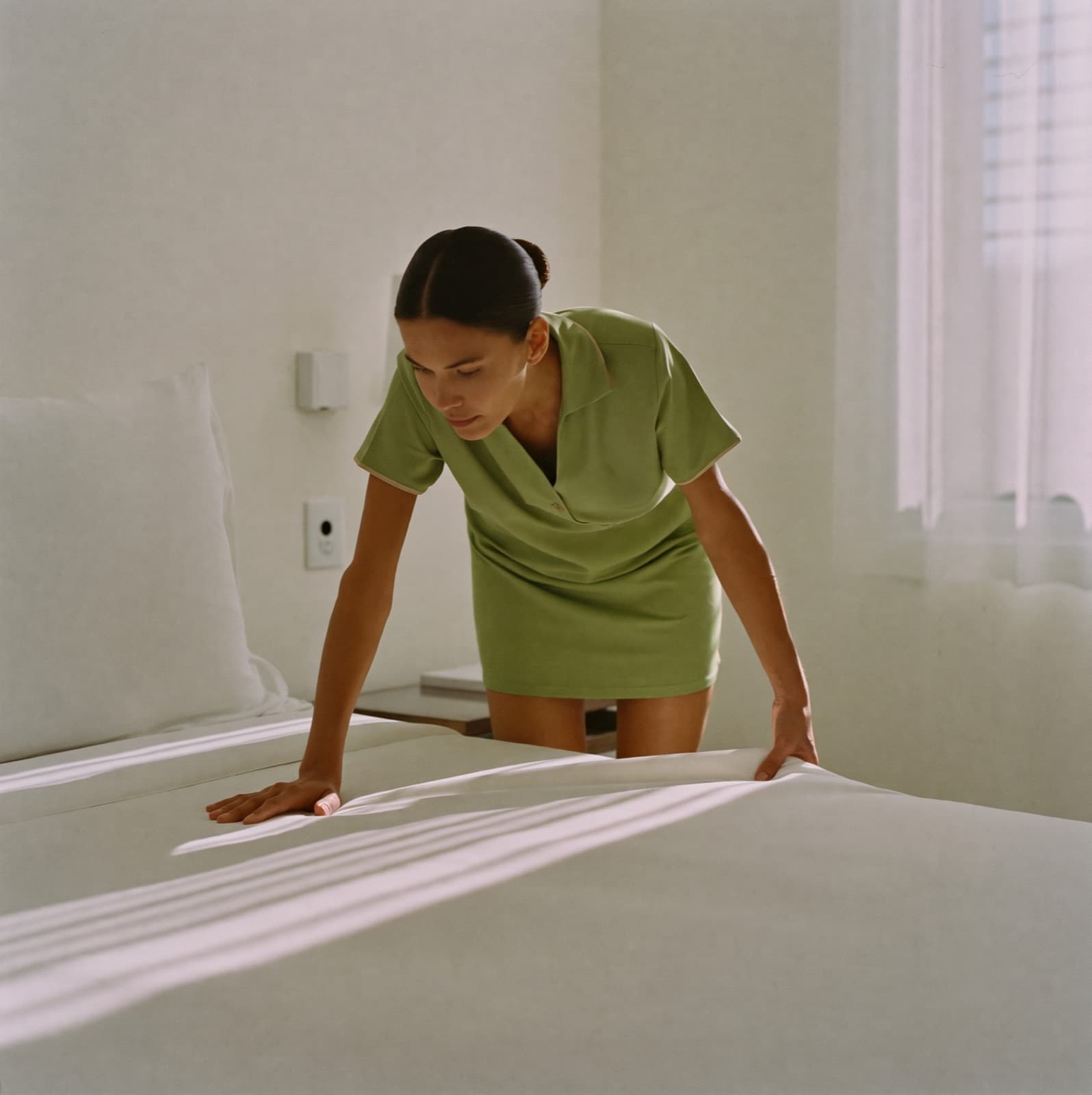 Una camarista alisa las sábanas de una cama con luz de la mañana.